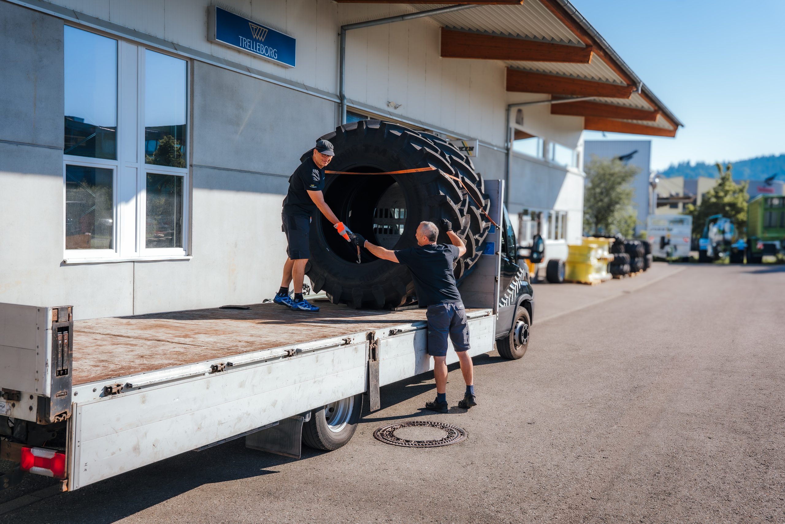 Zwei Mitarbeiter verladen einen großen Traktorreifen auf eine Pritsche vor der Halle