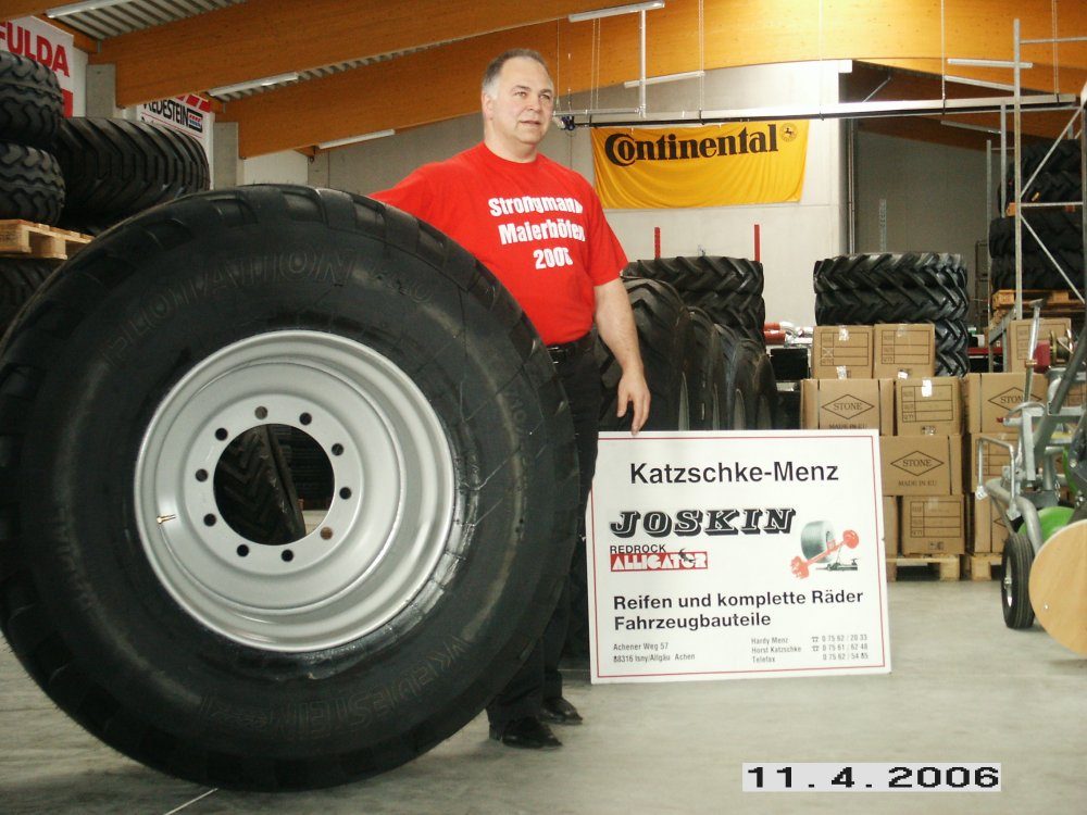 Mann im Strongman-Shirt neben einem großen Reifen mit Felge und dem Schild Katzschke-Menz